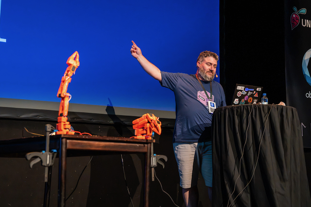 James on stage at Goatmire 2026, pointing upwards, with two orange SO-101 robot arms on a table beside him